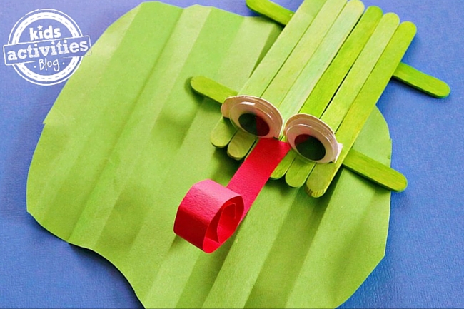 popsicle stick frog craft on a paper lily pad with a curly red tongue, googly eyes, and is green.
