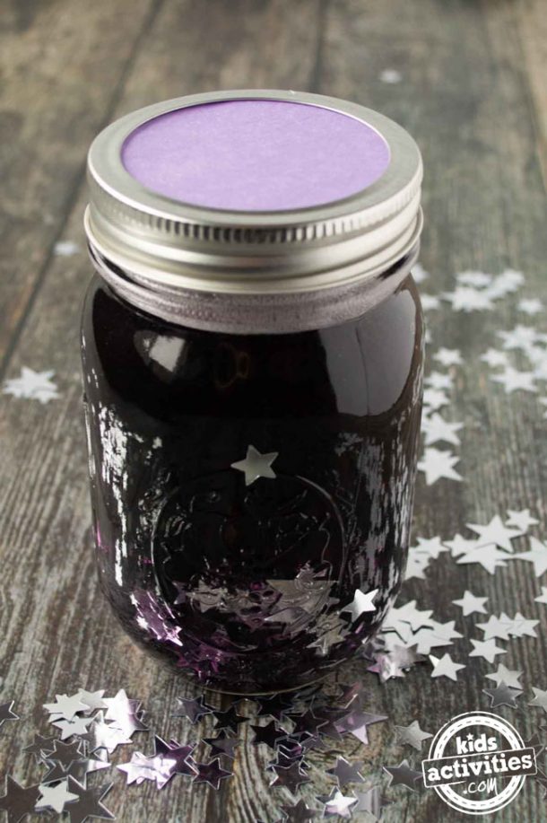 A mason jar filled with glue, purple water, and silver confetti with the top on. You can see purple construction paper under the lid. 