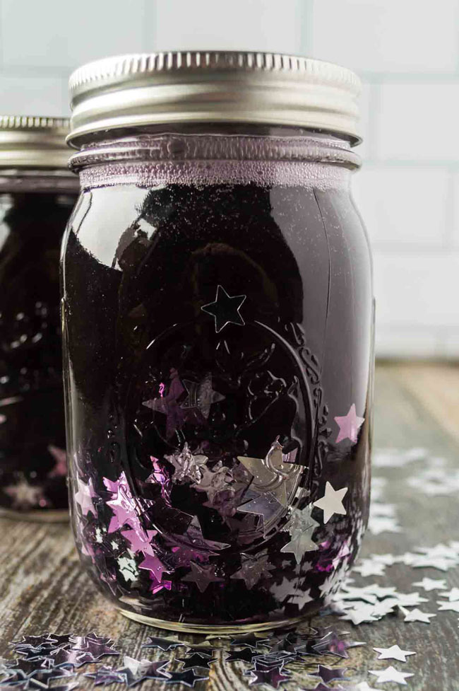 Falling stars glitter jar for kids contains clear glue, purple water, and silver star confetti. There is a second mason jar in the background.