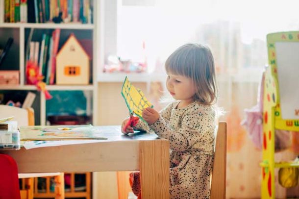 A preschool girl is cutting up a piece of yellow paper with her scissors while sitting at a small table in her room.