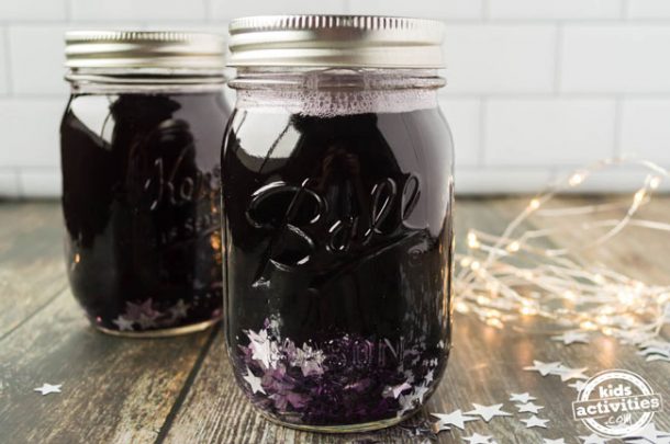 Two mason jars both filled with clear glue, purple water, and lots of silver confetti stars. There are star lights lit up in the background. 
