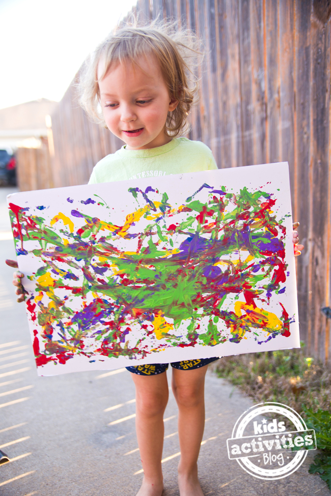 Painting With Balls- child holding up canvas of ball art with bright colors of orange, red, green, yellow and red