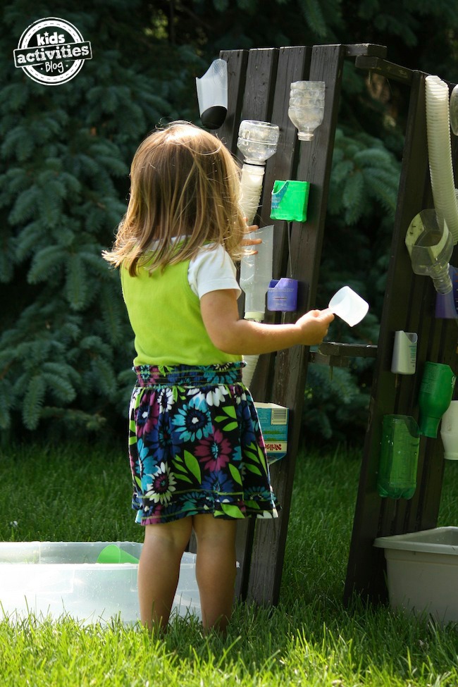 water wall for kids - water wall shown with multiple containers attached to wooden vertical surface fence and child with cup in hand