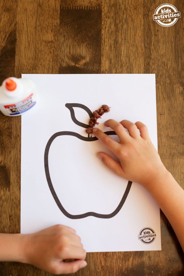 Apple printable on white cardstock with a boy's hand placing a ball of brown tissue paper on the apple's stem. Glue bottle sitting on the left side of the frame. 