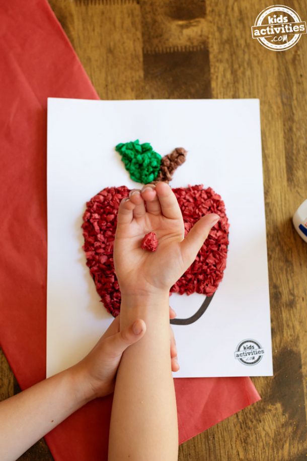 A boy holding a rolled up ball of red tissue paper above an in-progress tissue paper apple craft.