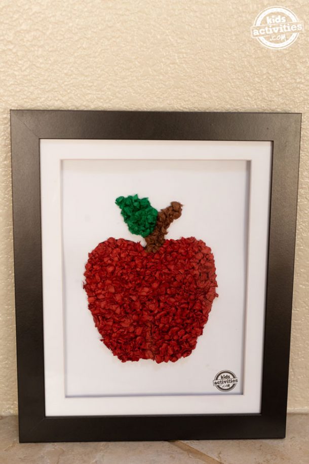 A black frame containing a piece of white cardstock with a red tissue paper apple on the paper.
