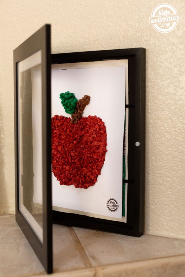 A black, front-loading frame halfway open to reveal a piece of white cardstock with a tissue paper apple on it. 