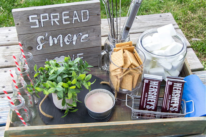 A wood box filled with ingredients to make s'mores including a handmade sign and bottles for drinks.