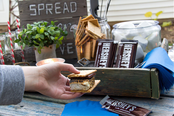 A hand holding s'mores in front of a backyard movie night s'mores kit. 