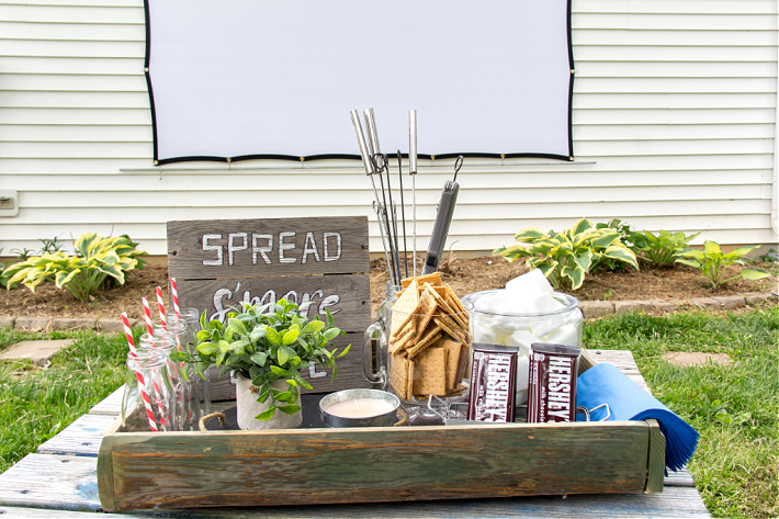 A box containing s'mores ingredients with an outdoor movie screen in the background for a backyard family movie night.