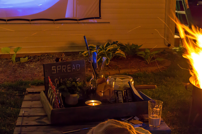 A s'mores kit on a table next to a fire pit while an outdoor movie plays on a screen in the background.
