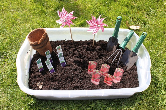 Plastic sensory bin full of dirt, seeds, and gardening tools on a patch of grass.