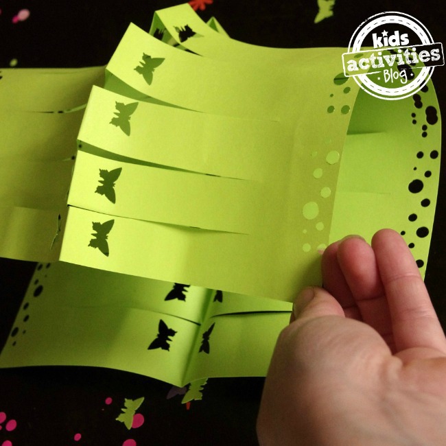How to use a paper punch to create butterflies and large and small dots on a green paper lantern that is being folded by a small hand.