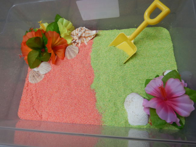Plastic bin full of edible green and pink sand with shells and tropical flowers and a yellow plastic shovel.