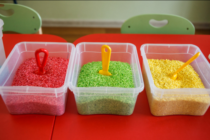 3 small sensory bins in plastic containers that are dry colored rice in red green and yellow