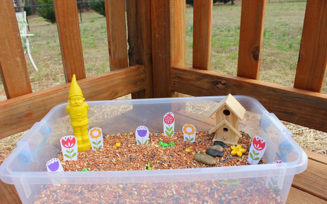 Plastic bin full of bird seed, a bird house, and various garden toys sitting on a wooden porch.