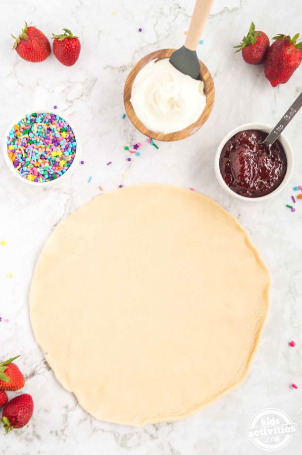 The four ingredients to make homemade pop tarts: pie crust rolled out, sprinkles, strawberry jam and vanilla frosting. 