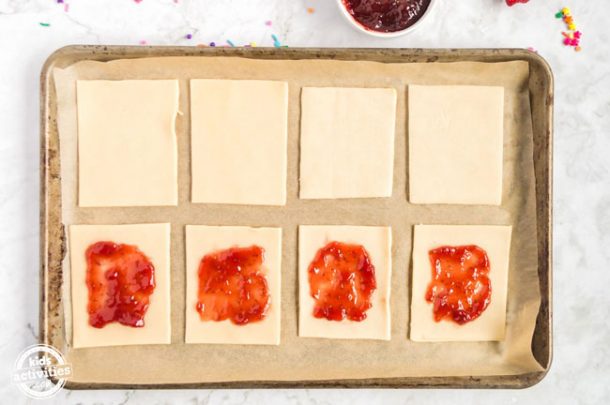 Eight rectangles of pie crust assembled on parchment paper on a baking sheet. Four of the pieces have strawberry jam on them. 