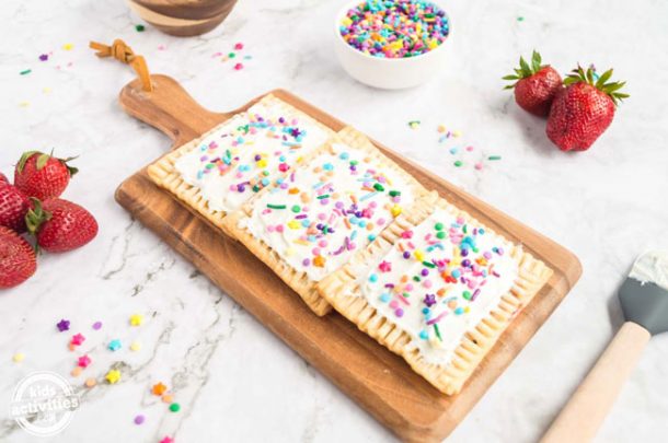 A wooden board containing three ready-to-eat homemade pop tarts, all covered in vanilla frosting and sprinkles. 