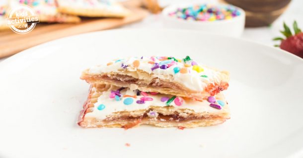 Two homemade pop tarts with vanilla frosting and sprinkles on top sitting on a white plate. More pop tarts and a bowl of sprinkles in the background.