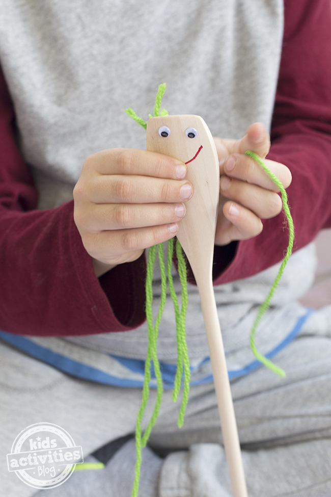 Spoon having yarn hair glued on, it is green, it has a red smiley face, and googly eyes.