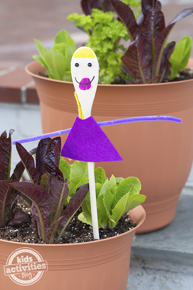 A finished wooden spoon craft with a pink dress, purple arms, pink lips, smiley face, googly eyes, and yellow hair.