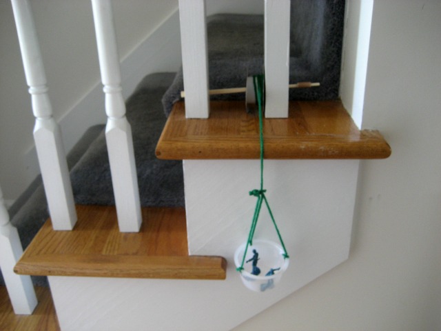 Homemade pulley close up - string wound around chopstick at the top with plastic basket raised from floor