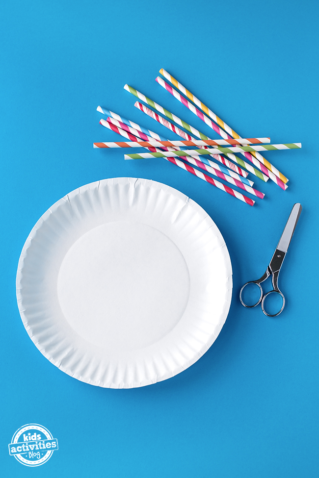 A paper plate, a pair of scissors, and some straws sit out on a blue background, ready to make a marble maze.