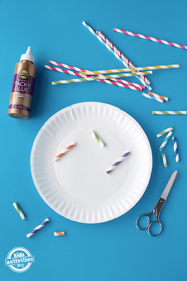 The scissors have been used to cut the straws into pieces. Tacky glue is now being used to attach the pieces of straw to the paper plate.